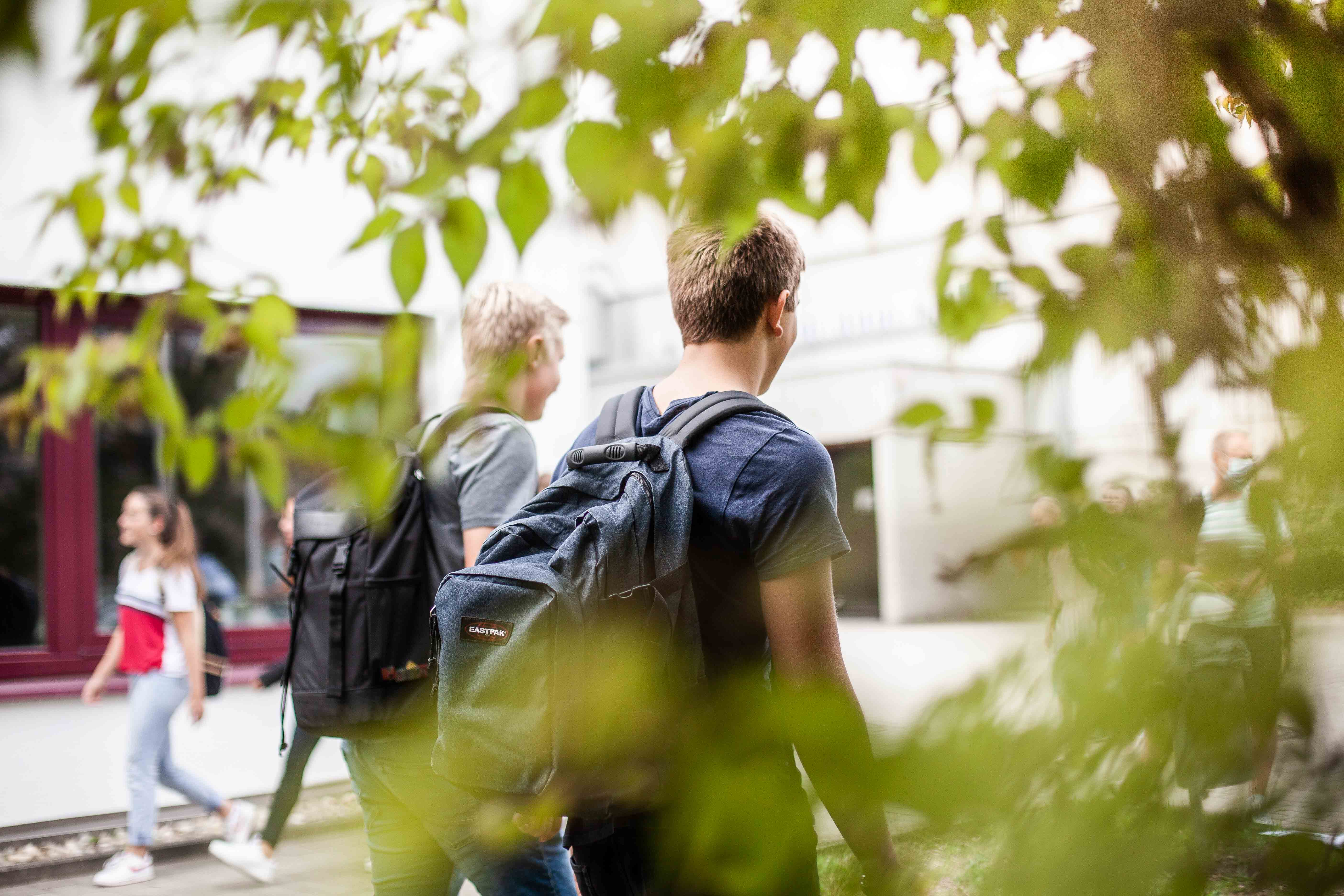 Zwei Schüler laufen nebeneinander mit Rucksäcken auf dem Pausenhof.